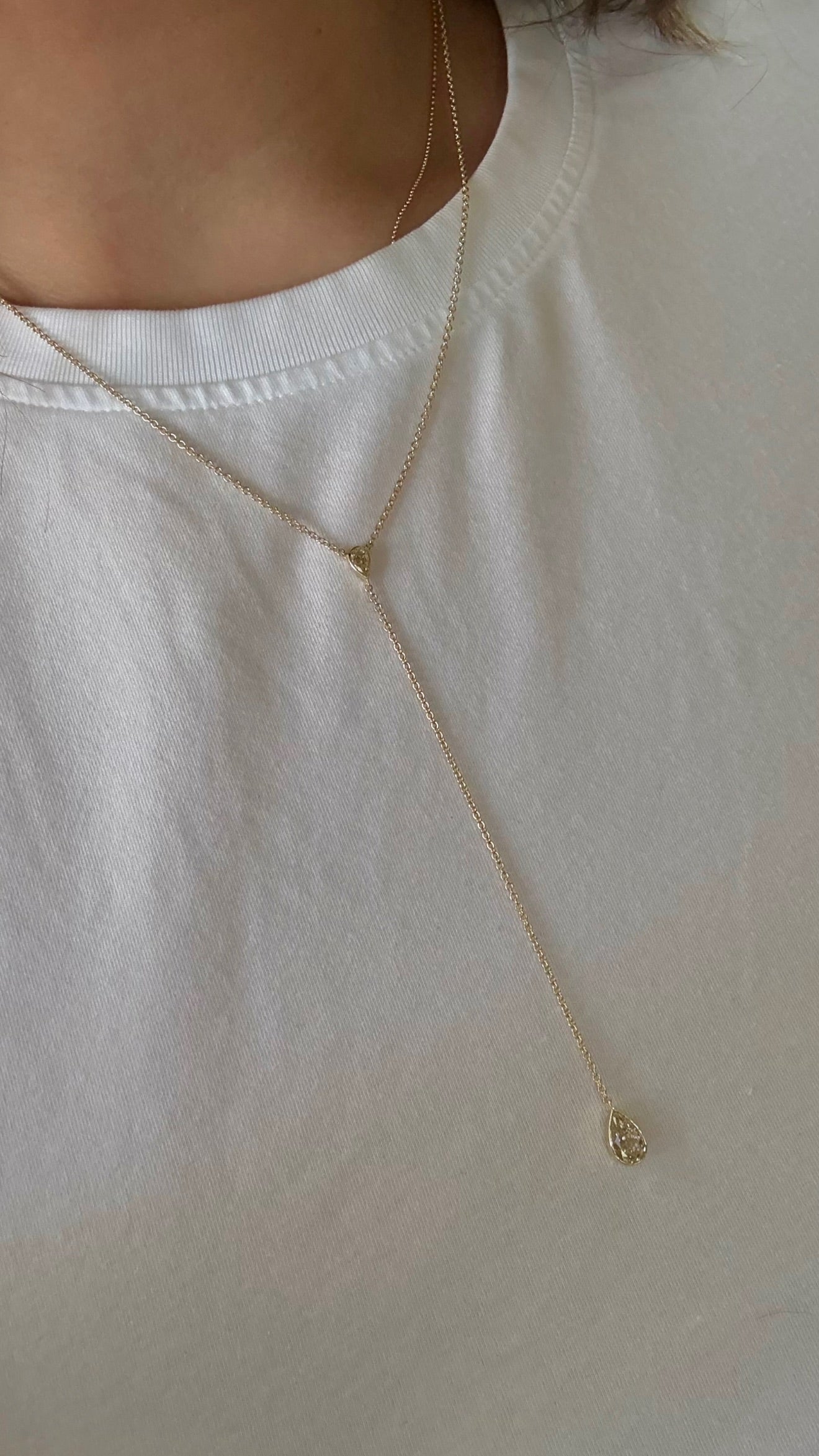 A close-up of a person wearing a delicate gold Y-shaped necklace with a small teardrop pendant over a white crew-neck t-shirt.