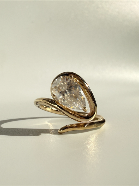 A gold ring featuring a large, teardrop-shaped clear gemstone in a prong setting, resting on a light gray surface with a soft shadow.